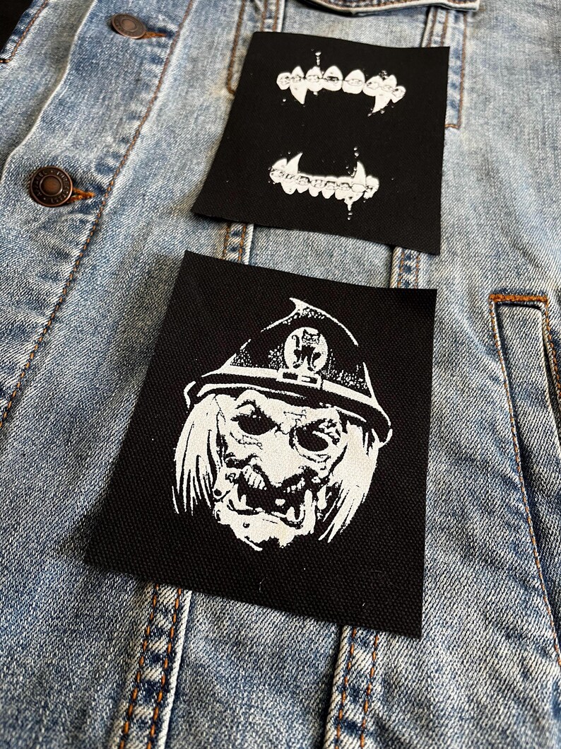 Peut inclure: Deux patchs thermocollants sur une veste en jean bleue. Un patch est noir avec une image blanche d'un monstre portant un chapeau et une grande bouche. L'autre patch est noir avec une image blanche d'une paire de crocs.