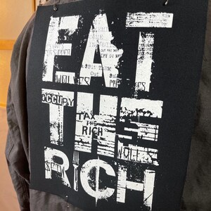 Pu&ograve; includere: Toppa in tessuto nero con testo bianco che recita "Eat the Rich" con altro testo che la circonda. Il testo &egrave; in uno stile usurato.