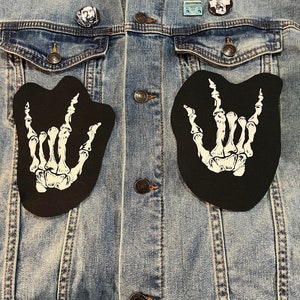 May include: A denim jacket with two skeleton hand patches, one on each side of the front. The patches are black with white bones and are in the shape of a hand making the rock and roll hand gesture. The jacket also has several pins, including a cassette tape, a person's face, and a skull.