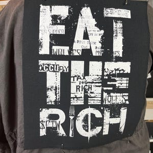 Pu&ograve; includere: Toppa nera e bianca con il testo "Eat the Rich" in un font usurato. La toppa ha una grafica di forchetta e simbolo del dollaro.