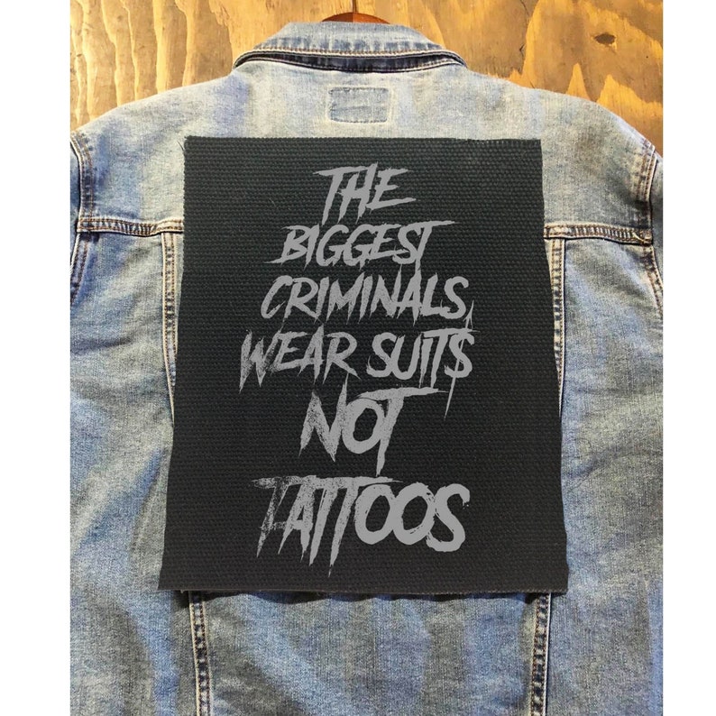 May include: A rectangular black fabric patch featuring the grey text THE BIGGEST CRIMINALS WEAR SUITS NOT TATTOOS in a distressed, jagged font. The patch is centered on the back of a light blue denim jacket with visible stitching and seams.