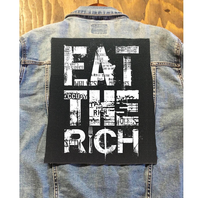 Pu&ograve; includere: Una toppa nera con testo bianco che recita "Eat the Rich" su una giacca di jeans. Il testo &egrave; in un font usurato e include altre parole come "Occupy" e "Tax the Rich".
