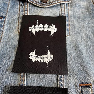 Peut inclure: Deux patchs thermocollants noirs avec des contours blancs de crocs. Les patchs sont sur une veste en jean bleu clair.