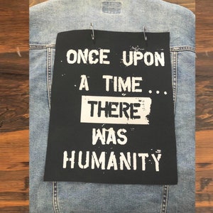 May include: A light blue denim jacket featuring a rectangular black fabric patch attached with two silver safety pins. The patch displays the white printed text: ONCE UPON A TIME... THERE WAS HUMANITY, with the word THERE enclosed in a white box.