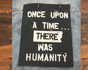May include: A light blue denim jacket featuring a rectangular black fabric patch attached with two silver safety pins. The patch displays the white printed text: ONCE UPON A TIME... THERE WAS HUMANITY, with the word THERE inside a white box.