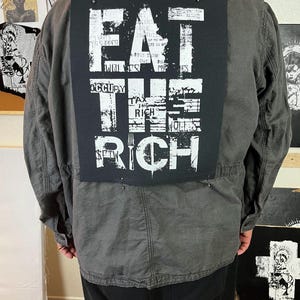 Pu&ograve; includere: Una giacca nera con una toppa bianca sul retro che dice "EAT THE RICH" con una grafica di una forchetta. La toppa &egrave; fatta di tessuto e ha un aspetto usurato.