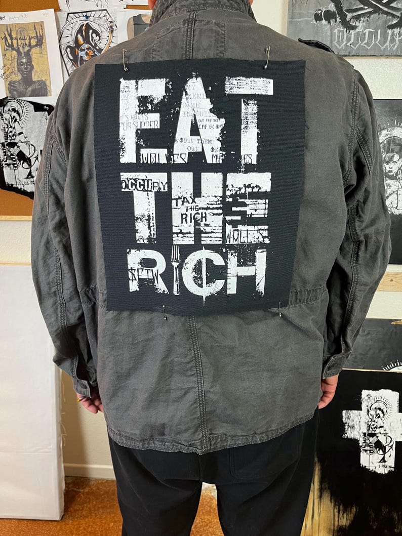 Pu&ograve; includere: Una giacca nera con una toppa bianca sul retro che dice "EAT THE RICH" con una grafica di una forchetta. La toppa &egrave; fatta di tessuto e ha un aspetto usurato.