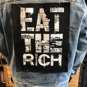 Pu&ograve; includere: Una giacca di jeans con una toppa nera sul retro che dice "EAT THE RICH" in lettere bianche. La toppa &egrave; realizzata in un tessuto strutturato e ha un aspetto usurato.