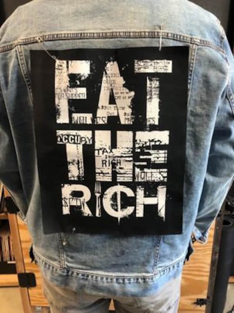 Pu&ograve; includere: Una giacca di jeans con una toppa nera sul retro che dice "EAT THE RICH" in lettere bianche. La toppa &egrave; realizzata in un tessuto strutturato e ha un aspetto usurato.