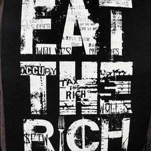 Pu&ograve; includere: Toppa bianca e nera con il testo "Eat the Rich" in un carattere audace e usurato. La toppa presenta una grafica di forchetta e dollaro. Sono visibili anche i testi "Occupy" e "Tax the Rich".