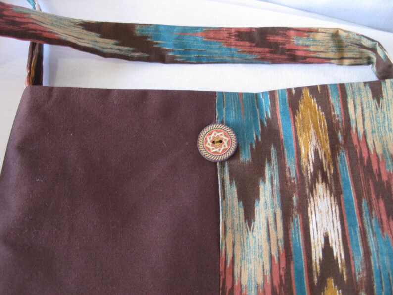 K&ouml;nnte beinhalten: Eine braune und blaugr&uuml;ne Stofftasche mit einem dekorativen Knopfverschluss. Die Tasche hat einen braunen Stoffriemen und ein gemustertes Design.