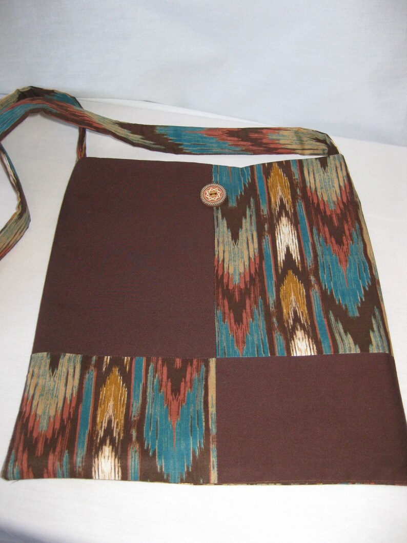 K&ouml;nnte beinhalten: Eine braune und t&uuml;rkisfarbene Patchwork-Tragetasche mit geometrischem Muster. Die Tasche hat einen braunen Trageriemen und einen dekorativen Knopf.