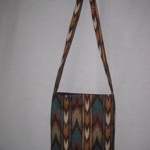 K&ouml;nnte beinhalten: Eine braune und blaue Schultertasche mit einem langen Trageriemen. Die Tasche hat ein geometrisches Muster in den Farben Braun, Blau, Wei&szlig; und Orange.