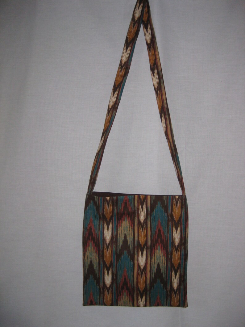 K&ouml;nnte beinhalten: Eine braune und blaue Schultertasche mit einem langen Trageriemen. Die Tasche hat ein geometrisches Muster in den Farben Braun, Blau, Wei&szlig; und Orange.