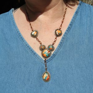 May include: A copper-toned necklace featuring five circular and heart-shaped settings containing multi-colored stone inlays in shades of orange, blue, and white. The necklace includes a central teardrop-shaped pendant suspended from a link chain.