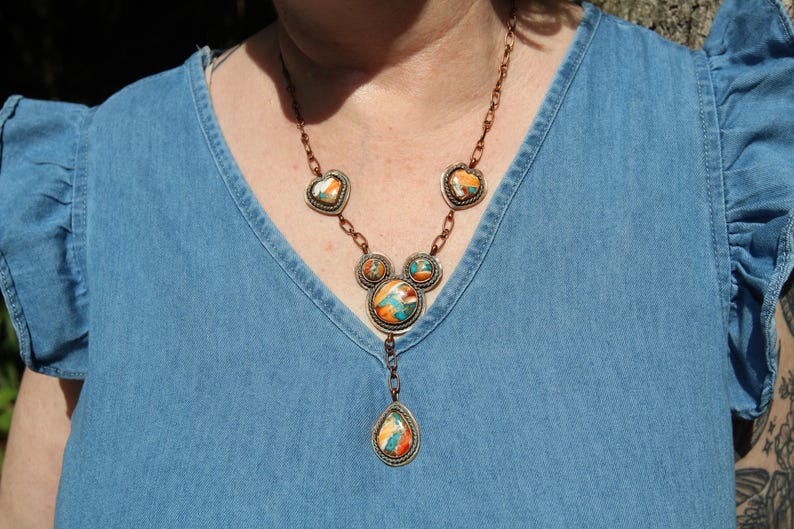 May include: A copper-toned necklace featuring five circular and heart-shaped settings containing multi-colored stone inlays in shades of orange, blue, and white. The necklace includes a central teardrop-shaped pendant suspended from a link chain.