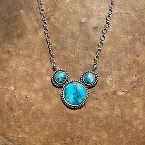 Pu&ograve; includere: Collana color argento con tre pietre turchesi. La pietra centrale &egrave; un grande turchese rotondo, affiancato da due pietre turchesi pi&ugrave; piccole. Le pietre sono incastonate in una lunetta argentata in stile corda e collegate da una delicata catena.