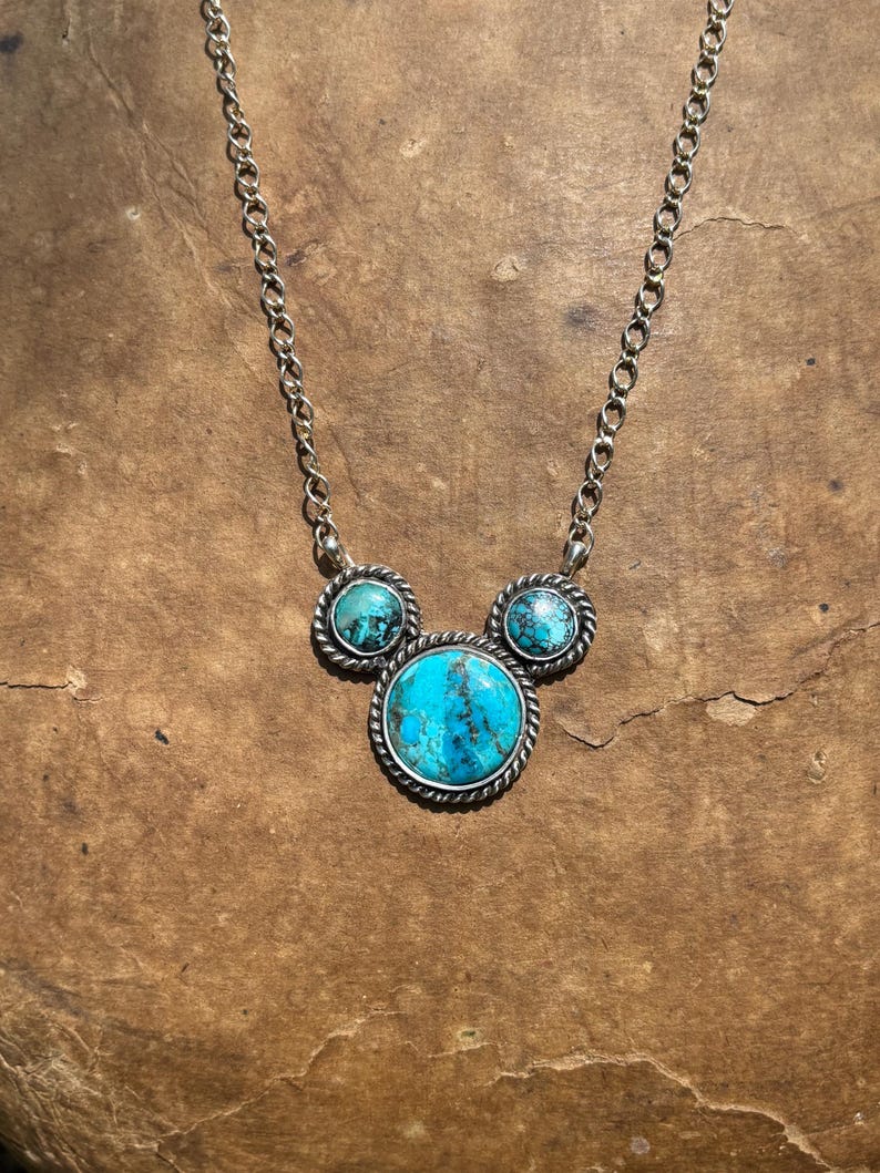 Pu&ograve; includere: Collana color argento con tre pietre turchesi. La pietra centrale &egrave; un grande turchese rotondo, affiancato da due pietre turchesi pi&ugrave; piccole. Le pietre sono incastonate in una lunetta argentata in stile corda e collegate da una delicata catena.