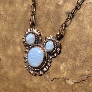May include: A necklace featuring three light blue, banded lace agate stones set in copper. The stones are surrounded by twisted wire and scalloped metal edges. The pendant is attached to a copper chain with elongated oval links.