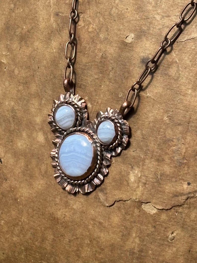May include: A necklace featuring three light blue, banded lace agate stones set in copper. The stones are surrounded by twisted wire and scalloped metal edges. The pendant is attached to a copper chain with elongated oval links.