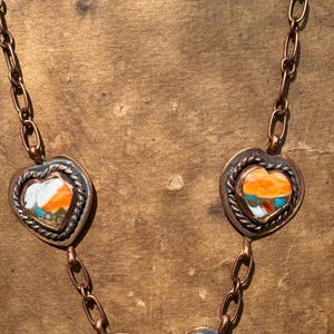 May include: A copper chain necklace featuring multiple heart-shaped and circular pendants. Each pendant is framed by a twisted copper border and contains inlaid stone segments in vibrant shades of orange, white, and turquoise blue.