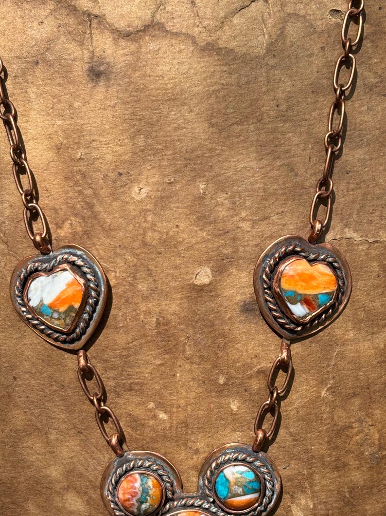 May include: A copper chain necklace featuring multiple heart-shaped and circular pendants. Each pendant is framed by a twisted copper border and contains inlaid stone segments in vibrant shades of orange, white, and turquoise blue.