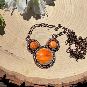 May include: A copper-toned necklace with a pendant featuring three orange-colored stones. The central stone is larger, surrounded by a twisted metal border. The necklace chain is also copper-toned. The background is a wooden surface with a white flower.