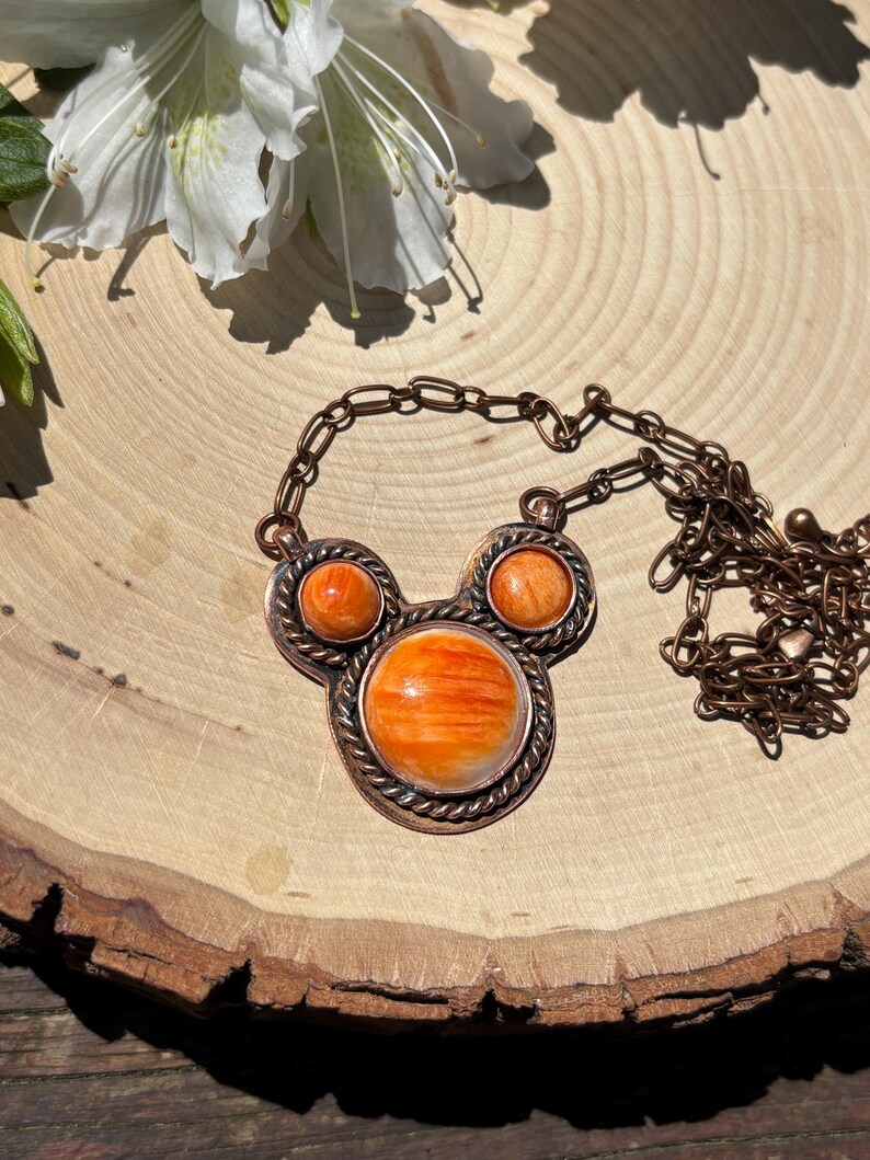 May include: A copper-toned necklace with a pendant featuring three orange-colored stones. The central stone is larger, surrounded by a twisted metal border. The necklace chain is also copper-toned. The background is a wooden surface with a white flower.