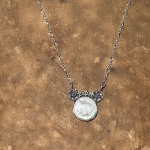 May include: A silver necklace featuring a round, iridescent white coin pearl pendant. The pendant is topped with a decorative silver element shaped like three small flowers. The chain consists of alternating oval and circular silver links.