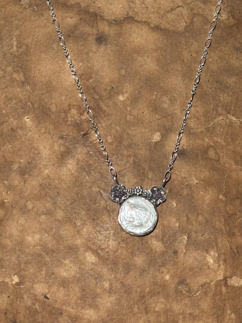 May include: A silver necklace featuring a round, iridescent white coin pearl pendant. The pendant is topped with a decorative silver element shaped like three small flowers. The chain consists of alternating oval and circular silver links.