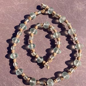 May include: A necklace made of light blue glass beads and brown leather cord. The beads are irregular in shape and have a slightly frosted finish.