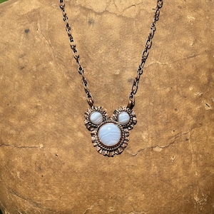 May include: A copper chain necklace featuring a pendant with three round, light blue banded agate stones set in scalloped copper bezels. The central stone is larger than the two smaller stones positioned above it, creating a cluster design.