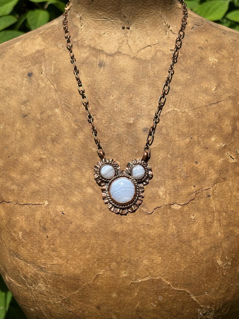 May include: A copper chain necklace featuring a pendant with three round, light blue banded agate stones set in scalloped copper bezels. The central stone is larger than the two smaller stones positioned above it, creating a cluster design.