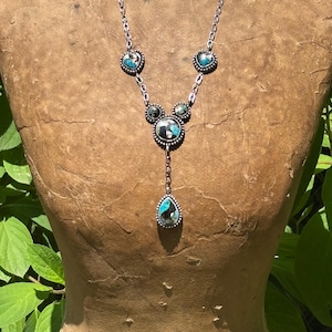 May include: A silver-toned necklace featuring a chain with four inlaid stone settings. Two heart-shaped stones, one circular stone, and one teardrop-shaped pendant hang from the chain. The stones display a mix of blue, black, and white patterns.
