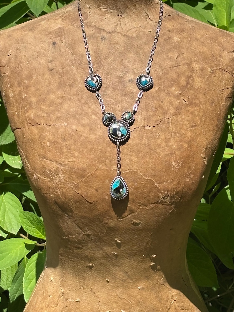 May include: A silver-toned necklace featuring a chain with four inlaid stone settings. Two heart-shaped stones, one circular stone, and one teardrop-shaped pendant hang from the chain. The stones display a mix of blue, black, and white patterns.