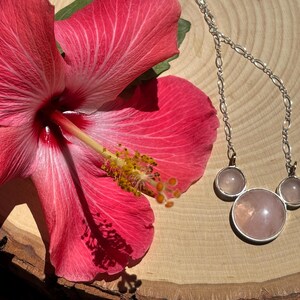 Puede incluir: Una vibrante flor de hibisco rosa junto a un collar de plata con tres colgantes de cuarzo rosa. El collar tiene una cadena delicada y los colgantes est&aacute;n engarzados en plata. La flor tiene un estambre amarillo.