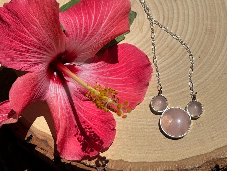 Puede incluir: Una vibrante flor de hibisco rosa junto a un collar de plata con tres colgantes de cuarzo rosa. El collar tiene una cadena delicada y los colgantes est&aacute;n engarzados en plata. La flor tiene un estambre amarillo.