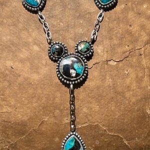 May include: A silver necklace featuring five inlaid stones with black, white, and turquoise blue patterns. The design includes two heart-shaped stones at the top, two smaller circular stones, a large central circular stone, and a teardrop pendant.