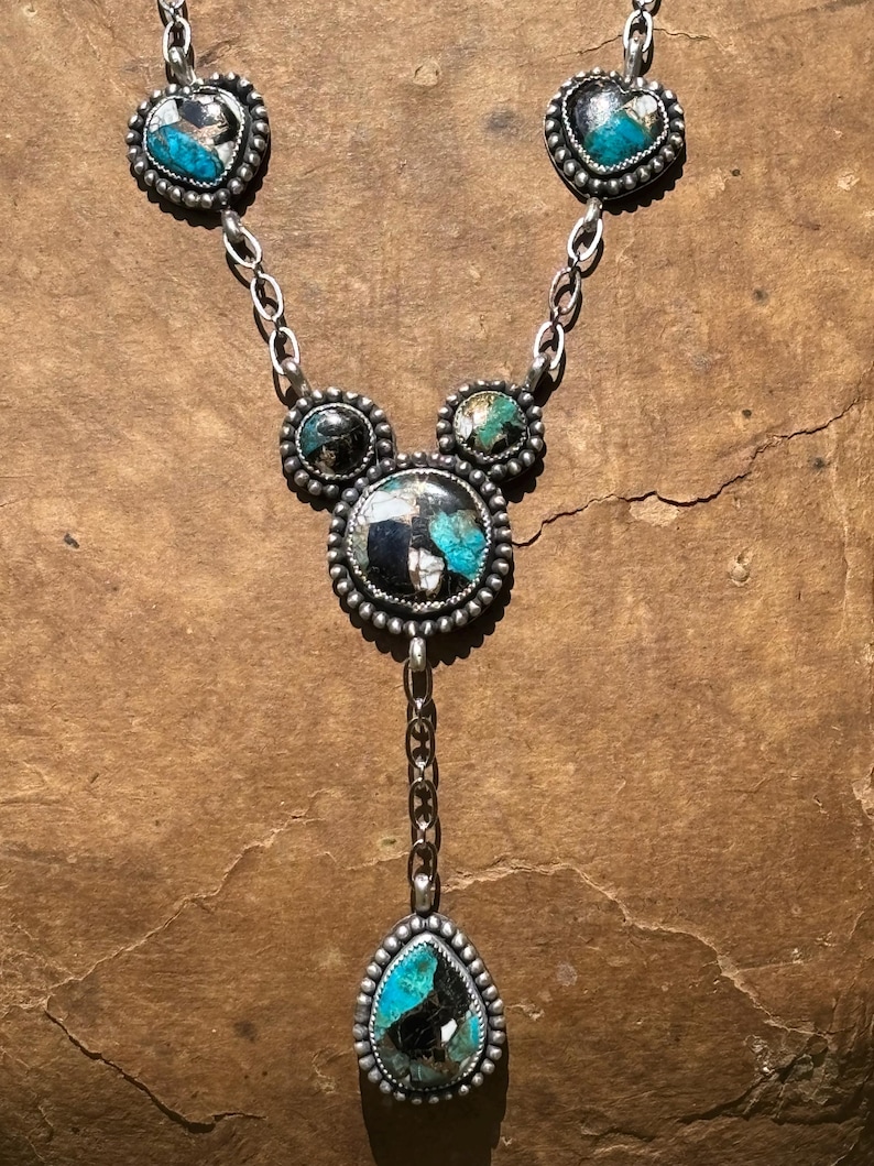 May include: A silver necklace featuring five inlaid stones with black, white, and turquoise blue patterns. The design includes two heart-shaped stones at the top, two smaller circular stones, a large central circular stone, and a teardrop pendant.