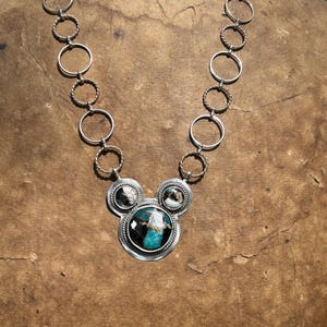 May include: A silver necklace with a pendant showcasing a large, round turquoise stone with black and white detailing. The pendant is framed by two smaller, circular stones. The chain is made of alternating rings and twisted rope-like sections.