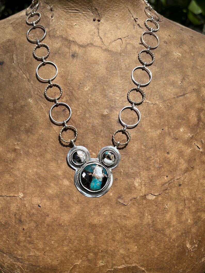 May include: A silver necklace with a pendant showcasing a large, round turquoise stone with black and white detailing. The pendant is framed by two smaller, circular stones. The chain is made of alternating rings and twisted rope-like sections.