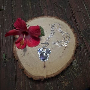 May include: A silver-toned necklace featuring a pendant with three circular, iridescent purple cabochons arranged in a cluster, suspended from a chain composed of interconnected silver rings, accompanied by a vibrant red hibiscus flower.