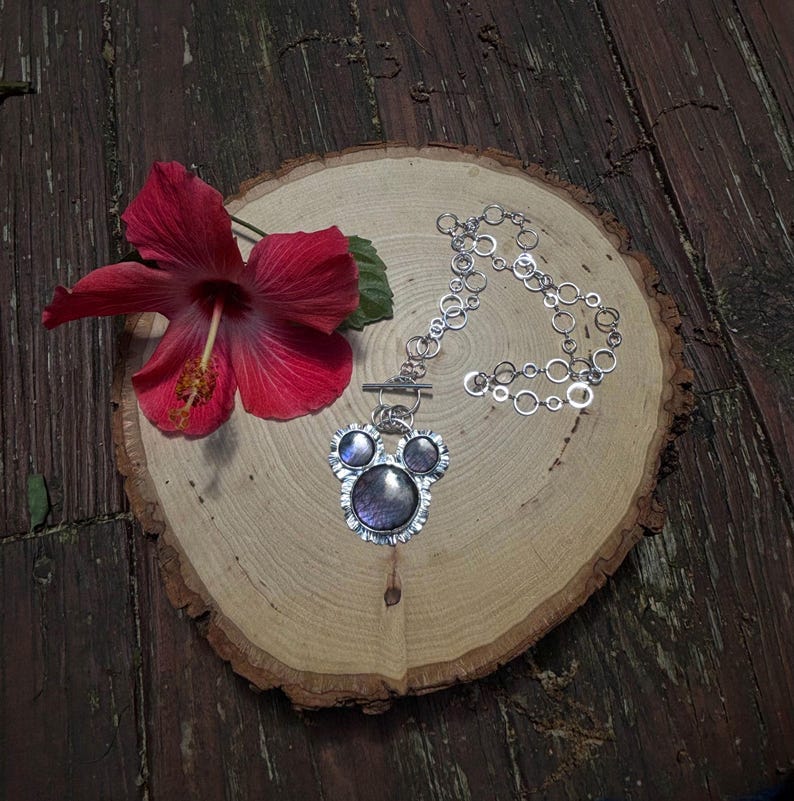 May include: A silver-toned necklace featuring a pendant with three circular, iridescent purple cabochons arranged in a cluster, suspended from a chain composed of interconnected silver rings, accompanied by a vibrant red hibiscus flower.