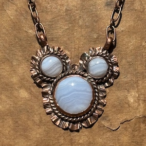 May include: A necklace featuring three round, light blue, banded agate cabochons set in copper. The central stone is the largest, flanked by two smaller stones. Each stone is framed by a twisted copper wire and a ruffled copper border.