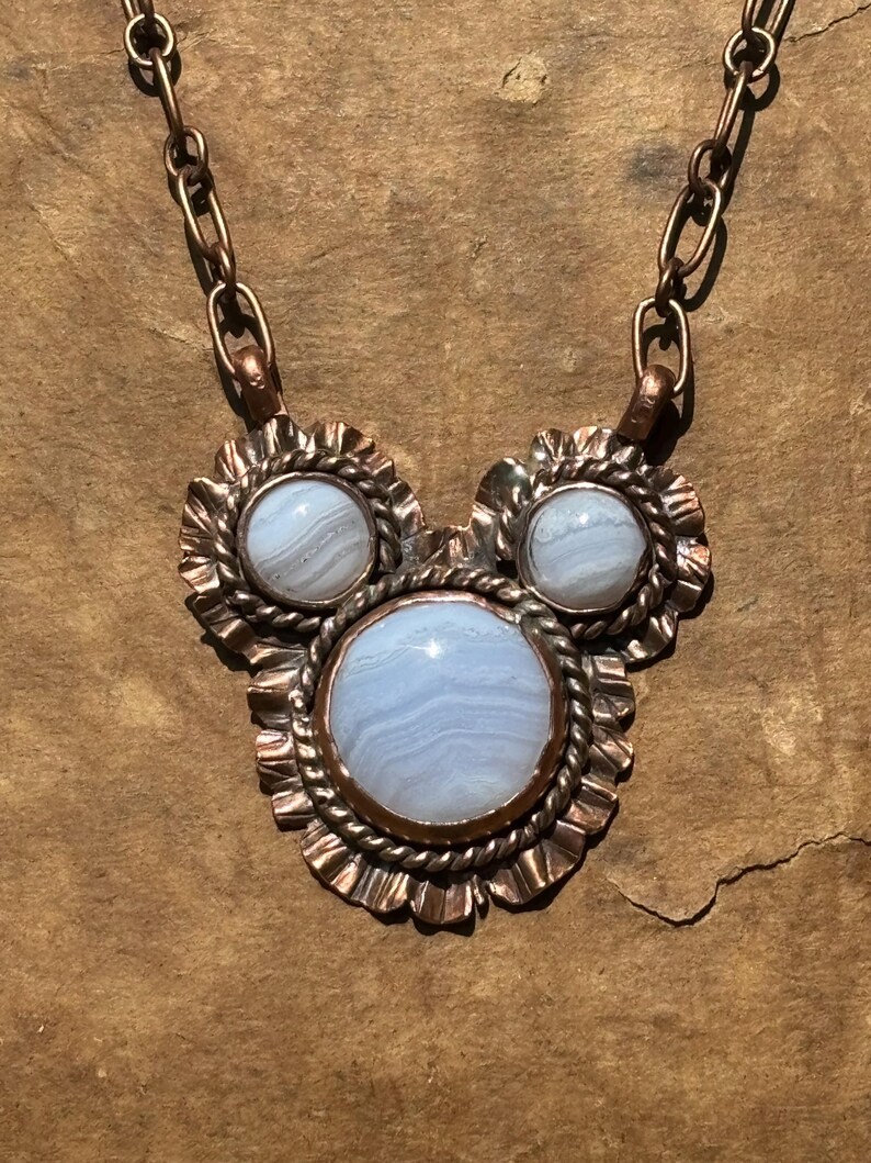 May include: A necklace featuring three round, light blue, banded agate cabochons set in copper. The central stone is the largest, flanked by two smaller stones. Each stone is framed by a twisted copper wire and a ruffled copper border.