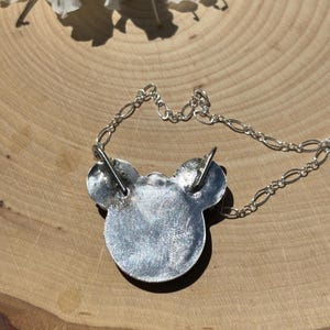 K&ouml;nnte beinhalten: Ein silberfarbener Metallanh&auml;nger in Form eines stilisierten Mauskopfes mit zwei runden Ohren. Der Anh&auml;nger hat eine texturierte, metallische Oberfl&auml;che und ist an einer feinen silberfarbenen Kette mit ovalen Gliedern befestigt, die oben an den Ohren ansetzt.