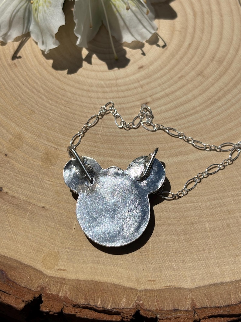 K&ouml;nnte beinhalten: Ein silberfarbener Metallanh&auml;nger in Form eines stilisierten Mauskopfes mit zwei runden Ohren. Der Anh&auml;nger hat eine texturierte, metallische Oberfl&auml;che und ist an einer feinen silberfarbenen Kette mit ovalen Gliedern befestigt, die oben an den Ohren ansetzt.