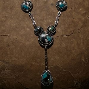 May include: A silver necklace featuring five bezel-set stones with turquoise and black matrix patterns. The design includes two heart-shaped stones, two small circular stones, one large central circular stone, and a teardrop-shaped pendant at the base.