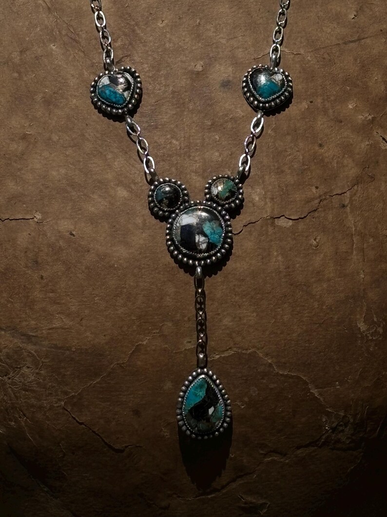 May include: A silver necklace featuring five bezel-set stones with turquoise and black matrix patterns. The design includes two heart-shaped stones, two small circular stones, one large central circular stone, and a teardrop-shaped pendant at the base.