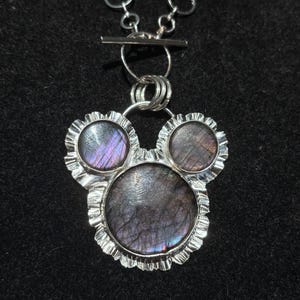 May include: A silver necklace featuring a pendant with three circular labradorite stones set in a ruffled metal frame. The stones display iridescent purple and grey tones. The pendant hangs from a silver chain with a toggle clasp closure.